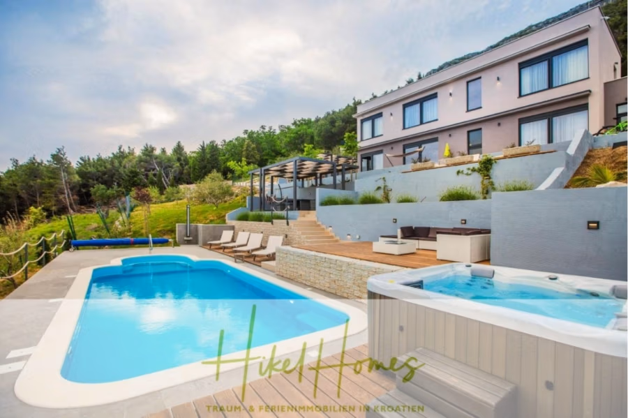Meerblick auf 2903m² großem Grundstück in wunderbarer Alleinlage am Stadtrand von Split - Eine moderne Villa am Hang mit großen Fenstern und Blick auf einen Swimmingpool und einen Whirlpool auf einer Holzterrasse. Der Bereich ist von üppigem Grün, Liegestühlen und einer Pergola umgeben, mit Bergen im Hintergrund. Unten das „Hikel Homes“-Logo. (41444)