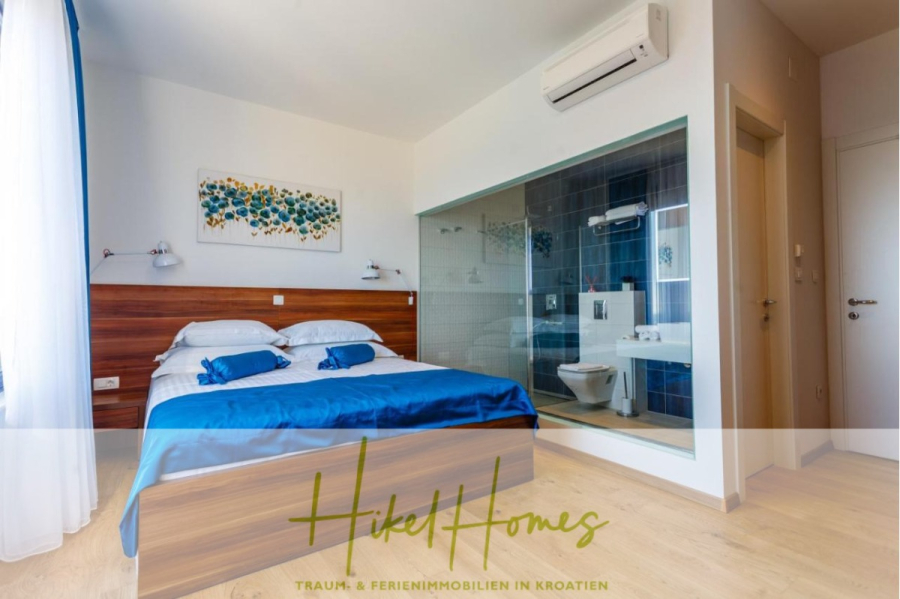 Meerblick auf 2903m² großem Grundstück in wunderbarer Alleinlage am Stadtrand von Split - Modernes Hotelzimmer mit einem Doppelbett, blauer und weißer Bettwäsche, Holzakzenten, einem Badezimmer mit Glaswänden und einer Klimaanlage über der Tür. Durch ein Fenster mit weißen Vorhängen strömt Sonnenlicht herein. (45845)