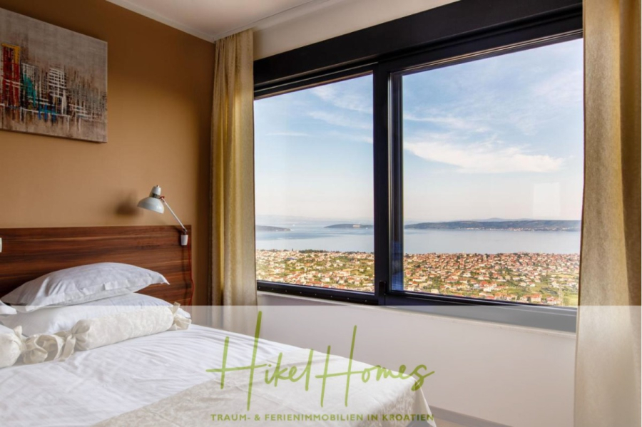 Meerblick auf 2903m² großem Grundstück in wunderbarer Alleinlage am Stadtrand von Split - Modernes Schlafzimmer mit einem ordentlich gemachten Bett, einem großen Fenster mit Blick auf eine Küstenstadt und das blaue Meer, beigen Vorhängen und einem Stadtbild an der Wand. Textüberlagerung liest "Hike Homes". (45850)