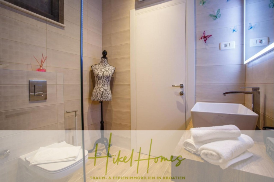 Meerblick auf 2903m² großem Grundstück in wunderbarer Alleinlage am Stadtrand von Split - Modernes Badezimmer mit beigen Fliesen, einer weißen Toilette, einem Gefäßwaschbecken, gefalteten Handtüchern, einer dekorativen Schaufensterpuppe und bunten Schmetterlings-Wanddekorationen. Der Raum sieht sauber und einladend aus und verfügt über ausreichend Licht. (45838)