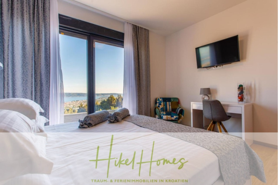 Meerblick auf 2903m² großem Grundstück in wunderbarer Alleinlage am Stadtrand von Split - Helles Schlafzimmer mit einem sauber gemachten Doppelbett, einem großen Fenster mit Blick aufs Meer, einem Schreibtisch mit Stuhl, einem an der Wand montierten Fernseher und hellem Dekor. Der Text am unteren Rand lautet "Hikel Homes". (45851)