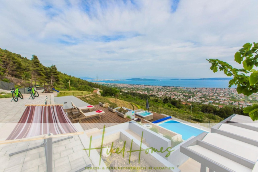 Meerblick auf 2903m² großem Grundstück in wunderbarer Alleinlage am Stadtrand von Split - Moderne Villa in Hanglage mit Pool, Sonnenliegen und grünen Fahrrädern mit Blick auf eine Küstenstadt und das weite blaue Meer unter einem teilweise bewölkten Himmel, umgeben von üppigem Grün und fernen Bergen. (45867)