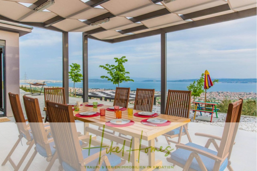 Meerblick auf 2903m² großem Grundstück in wunderbarer Alleinlage am Stadtrand von Split - Essbereich im Freien mit einem Holztisch für acht Personen unter einer modernen Pergola. Die Terrasse bietet einen malerischen Blick auf die Küste, das blaue Meer, die Stadt und die fernen Hügel. Kleine Bäume und bunte Liegestühle stehen in der Nähe. (45863)