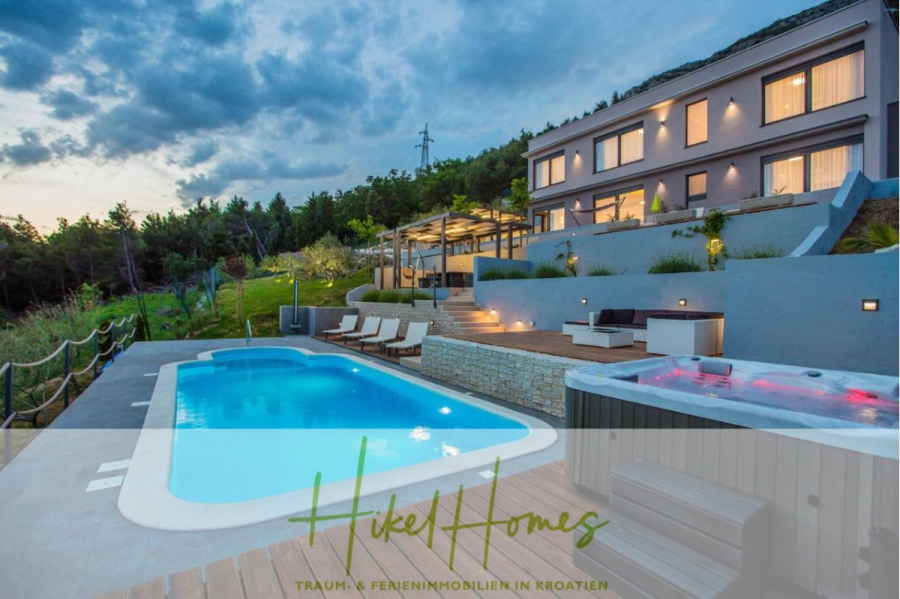 Meerblick auf 2903m² großem Grundstück in wunderbarer Alleinlage am Stadtrand von Split - Moderne Villa in den Bergen in der Abenddämmerung mit beleuchteten Fenstern, einem Außenpool, einem Whirlpool, Liegestühlen und umgeben von Grünflächen. Das Logo und der Slogan von "Hikel Homes" sind am unteren Rand des Bildes eingeblendet. (45862)