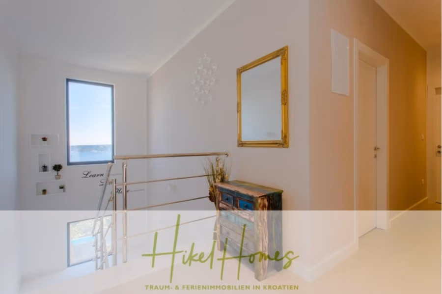 Meerblick auf 2903m² großem Grundstück in wunderbarer Alleinlage am Stadtrand von Split - Heller Flur mit großem Fenster und Meerblick. Links ein Metallgeländer, rechts eine beige Wand mit gerahmtem Spiegel und ein kleiner rustikaler Schrank mit bunter Front. Unten steht „Hikel Homes“. (41430)