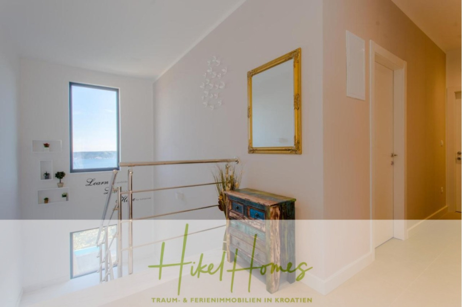 Meerblick auf 2903m² großem Grundstück in wunderbarer Alleinlage am Stadtrand von Split - Ein heller Flur mit beigen Wänden, einer Holzkommode mit blauen Schubladen, einem goldgerahmten Spiegel und einem Treppengeländer aus Metall. Ein großes Fenster lässt natürliches Licht herein. Das Logo von "Hikel Homes" und kroatischer Text überlagern das Bild. (45841)
