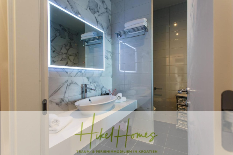 Meerblick auf 2903m² großem Grundstück in wunderbarer Alleinlage am Stadtrand von Split - Modernes Badezimmer mit einem leuchtenden rechteckigen Spiegel, einer Marmorwand, einem Gefäßwaschbecken auf einer weißen Arbeitsplatte, Handtüchern auf einem Regal und grau gekachelten Wänden. Der Text "Hikel Homes" erscheint am unteren Rand. (45854)