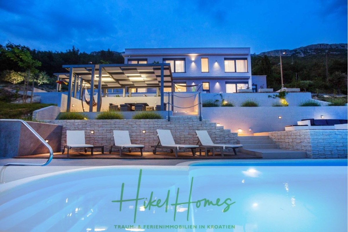 Moderne Villa mit großen Fenstern und Außenbeleuchtung, Sitzgelegenheiten auf der Terrasse und Liegestühlen am Pool, umgeben von einem beleuchteten Pool in der Abenddämmerung und einer Gartenanlage. Der Text lautet: "Hikel Homes - Traum & Ferienimmobilien in Kroatien. - Meerblick auf 2903m² großem Grundstück in wunderbarer Alleinlage am Stadtrand von Split