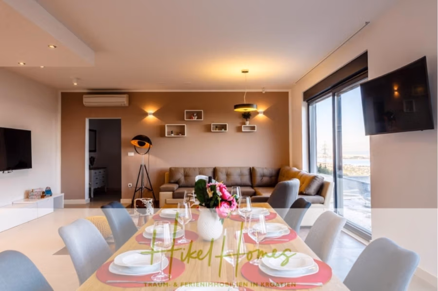 Meerblick auf 2903m² großem Grundstück in wunderbarer Alleinlage am Stadtrand von Split - Ein moderner Wohn- und Essbereich mit einem großen beigefarbenen Sofa, einer stilvollen Stehlampe und einem Wandfernseher. Der Esstisch ist mit rosa Tellern und Gläsern gedeckt. Große Fenster bieten einen malerischen Ausblick und sorgen für natürliches Licht im offenen Raum. (41432)