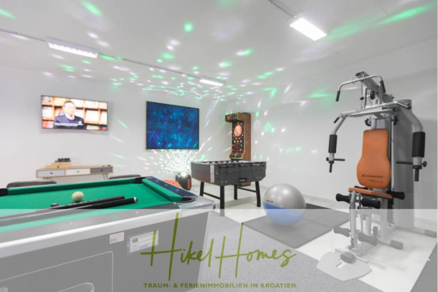 Meerblick auf 2903m² großem Grundstück in wunderbarer Alleinlage am Stadtrand von Split - Ein modernes Spielzimmer mit Billardtisch, Tischfußball, Dartscheibe, Fitnessgeräten, Fernseher an der Wand und bunten Discolichtern. Auf dem Boden steht "Hikel Homes Traum- & Ferienimmobilien in Kroatien". (45857)