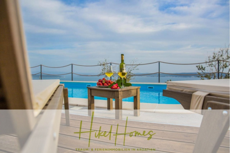 Meerblick auf 2903m² großem Grundstück in wunderbarer Alleinlage am Stadtrand von Split - Auf einem Tisch am Pool stehen eine Flasche Wein, zwei Gläser und ein Teller mit Erdbeeren, umgeben von Liegestühlen mit Blick auf das Meer und den Himmel im Hintergrund. Der Schriftzug "Hikel Homes" erscheint am unteren Rand. (45871)