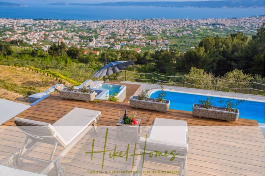 Meerblick auf 2903m² großem Grundstück in wunderbarer Alleinlage am Stadtrand von Split - Ein malerischer Blick auf die Hügellandschaft mit einer Poolterrasse mit Liegestühlen und einem kleinen Tisch. Darunter erstreckt sich eine Küstenstadt mit üppiger Landschaft und blauem Wasser in der Ferne. Auf der Terrasse steht ein Sonnenschirm, und auf dem Tisch stehen eine Flasche mit Gläsern. (41455)