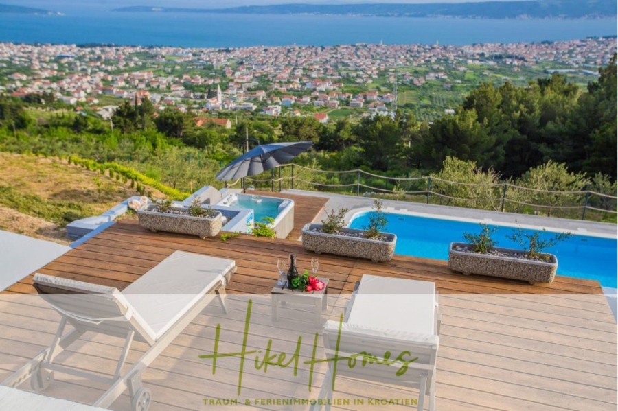 Meerblick auf 2903m² großem Grundstück in wunderbarer Alleinlage am Stadtrand von Split - Eine Holzterrasse mit Liegestühlen, einem Pool und einem Sonnenschirm bietet einen Blick auf ein üppiges Tal und eine Küstenstadt am Meer. Flaschen und Gläser stehen auf einem Tisch. Der Text lautet "Hikel Homes - Traum- & Ferienimmobilien in Kroatien. (45873)