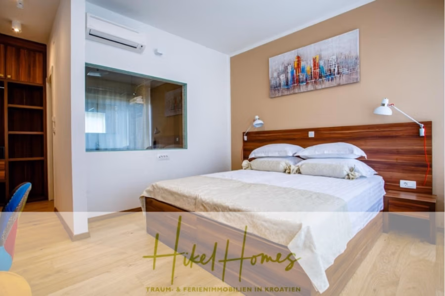 Meerblick auf 2903m² großem Grundstück in wunderbarer Alleinlage am Stadtrand von Split - Modernes Schlafzimmer mit einem hölzernen Doppelbett, frischer weißer Bettwäsche und passenden Nachttischlampen. Eine Glaswand trennt einen kleinen Bereich mit Regalen ab. Über der Glaswand befindet sich eine Klimaanlage, und ein farbenfrohes Stadtbild ziert die beige Wand. Der Boden ist aus hellem Holz. (41431)