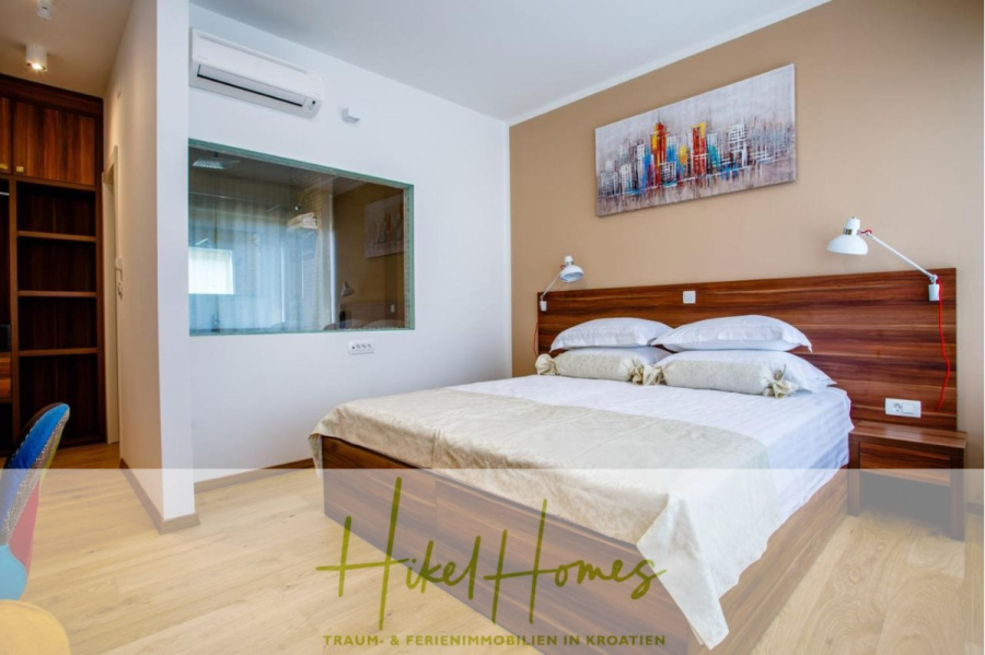 Meerblick auf 2903m² großem Grundstück in wunderbarer Alleinlage am Stadtrand von Split - Ein modernes Schlafzimmer mit einem Holzbett, weißer Bettwäsche, Wandkunst über dem Kopfteil, Nachttischlampen, einem Glasfenster zu einem anderen Raum, Klimaanlage und Holzfußboden. Die Textüberlagerung lautet "Hikel Homes". (45842)