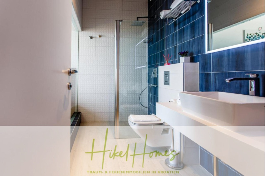 Meerblick auf 2903m² großem Grundstück in wunderbarer Alleinlage am Stadtrand von Split - Modernes Badezimmer mit einer Glasdusche, blau gefliester Wand, weißem Waschbecken und Toilette, großem Spiegel und Chromarmaturen. Der Logo-Text am unteren Rand lautet "Hikel Homes Traum- & Ferienimmobilien in Kroatien. (45847)