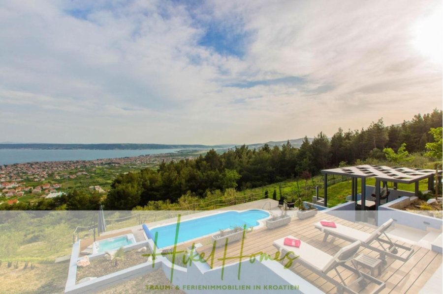 Meerblick auf 2903m² großem Grundstück in wunderbarer Alleinlage am Stadtrand von Split - Von der Terrasse am Hang mit Pool, Liegestühlen und Pergola überblickt man eine malerische Bucht, eine Küstenstadt und bewaldete Hügel unter einem teilweise bewölkten Himmel. (45866)