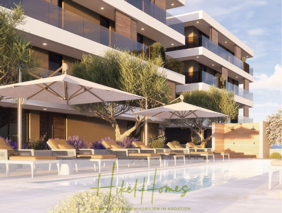 1. Reihe hochwertige Neubau-Meerblick Wohnung mit 2 SZ, Terrasse und Gemeinschaftspool - Ein modernes Apartmentgebäude mit Balkonen blickt auf einen Swimmingpool mit Sonnenliegen und großen Sonnenschirmen. Hohe Bäume und Sträucher umgeben den Poolbereich und schaffen eine ruhige und luxuriöse Atmosphäre unter einem klaren blauen Himmel. (42734)