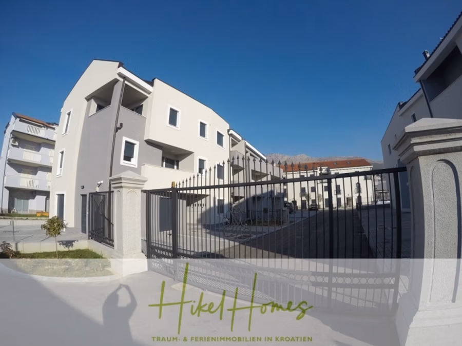 Galerie Wohnung, 3 Schlafzimmer / 70m zum Meer / Pool / 2 Bäder - Modernes Wohngebäude mit weiß-grauer Fassade, umgeben von einem schwarzen Metalltor. Die Szene ist sonnig und der Himmel klar. Unten ist das Logo von „Hikel Homes“ mit Text über Ferienimmobilien in Kroatien eingeblendet. (41262)