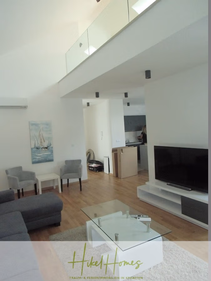 Galerie Wohnung, 3 Schlafzimmer / 70m zum Meer / Pool / 2 Bäder - Modernes Wohnzimmer mit hoher Decke und Balkon mit Glasgeländer. Es verfügt über einen Flachbildfernseher, graue Sofas und einen Couchtisch aus Glas. An der Wand hängt ein Gemälde eines Segelboots, im Hintergrund ist eine offene Küche zu sehen. (41256)