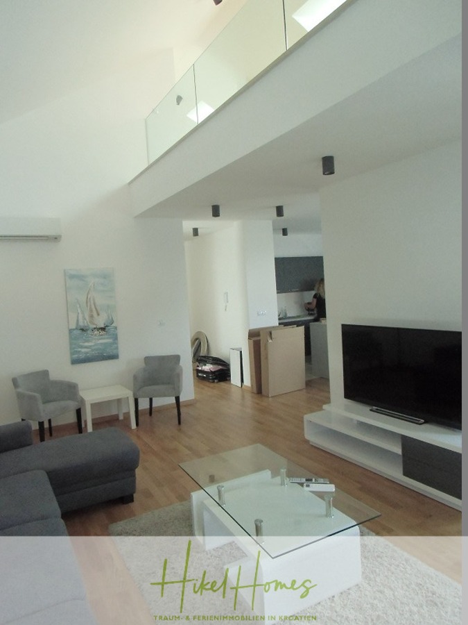 Galerie Wohnung, 3 Schlafzimmer / 70m zum Meer / Pool / 2 Bäder - Modernes Wohnzimmer mit grauen Möbeln, einem Couchtisch aus Glas, einem an der Wand montierten Fernseher, einem Gemälde eines Segelboots, Holzboden und einer offenen Küche im Hintergrund. Der Raum ist hell mit hohen Decken und weißen Wänden. (46577)