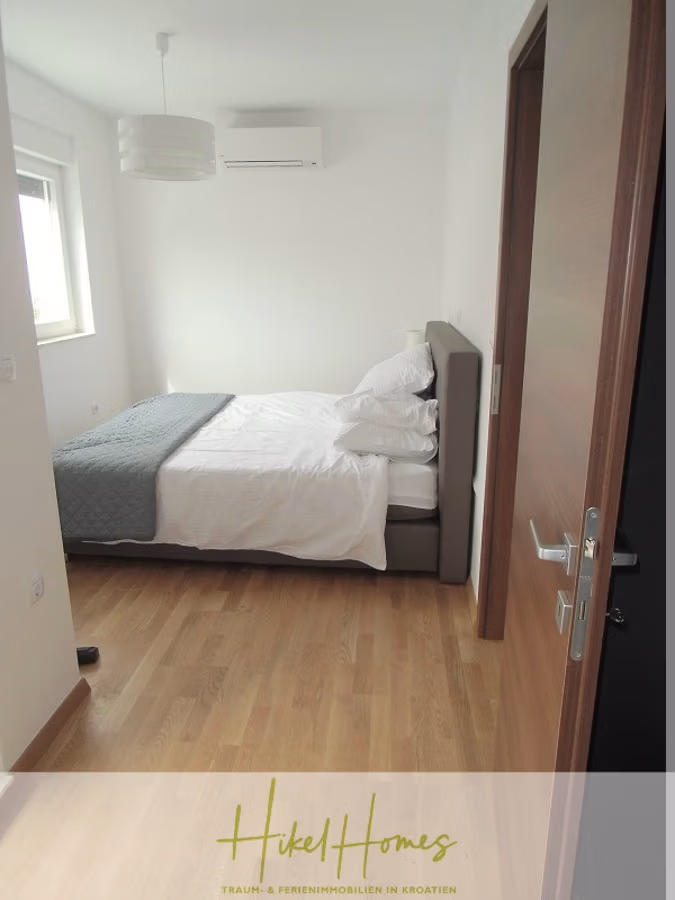 Galerie Wohnung, 3 Schlafzimmer / 70m zum Meer / Pool / 2 Bäder - Ein minimalistisches Schlafzimmer mit einem ordentlich gemachten Bett, weißer Bettwäsche und einer grauen Decke. Ein Fenster links lässt Tageslicht herein. Das Zimmer verfügt über Parkettboden, eine Deckenleuchte und eine wandmontierte Klimaanlage. Eine Holztür ist teilweise geöffnet. (41257)