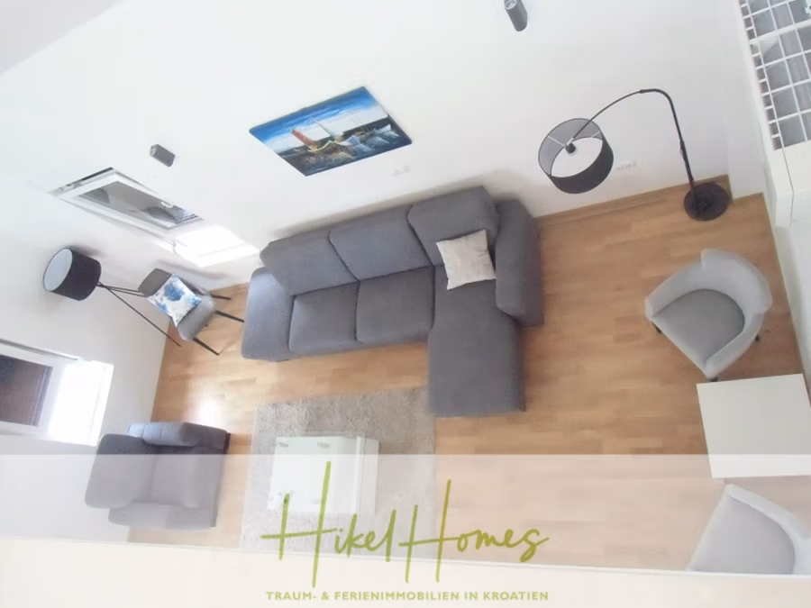 Galerie Wohnung, 3 Schlafzimmer / 70m zum Meer / Pool / 2 Bäder - Ein modernes Wohnzimmer mit Holzboden, einem grauen L-förmigen Sofa, einem grauen Sessel, einer Stehlampe und einem Couchtisch mit Glasplatte auf einem hellen Teppich. An der Wand hängt ein Gemälde. Unten steht der Text „Hikel Homes“. (41254)
