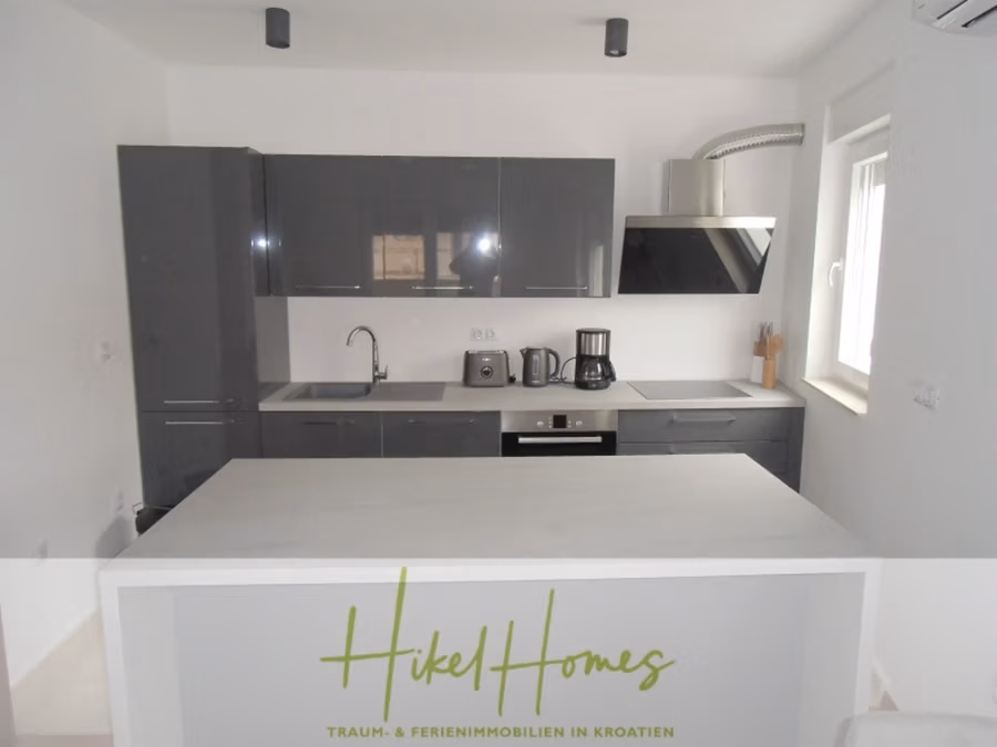 Galerie Wohnung, 3 Schlafzimmer / 70m zum Meer / Pool / 2 Bäder - Moderne Küche mit glänzenden grauen Schränken, einem eingebauten Backofen und einer eleganten Dunstabzugshaube. Zu den Küchengeräten gehören eine Kaffeemaschine und ein Wasserkocher. Im Vordergrund steht eine helle Kücheninsel. Ein Fenster rechts sorgt für Tageslicht. (41252)
