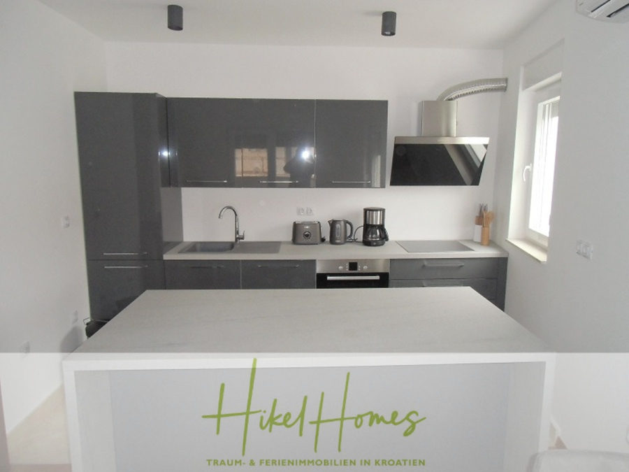 Galerie Wohnung, 3 Schlafzimmer / 70m zum Meer / Pool / 2 Bäder - Moderne Küche mit glänzenden grauen Schränken, eingebautem Backofen, Kaffeemaschine, Toaster, Wasserkocher, Spüle und Abzugshaube. Weiße Arbeitsplatte im Vordergrund. Ein Fenster auf der rechten Seite bringt natürliches Licht herein. Der Schriftzug "Hikel Homes" am unteren Rand. (46571)