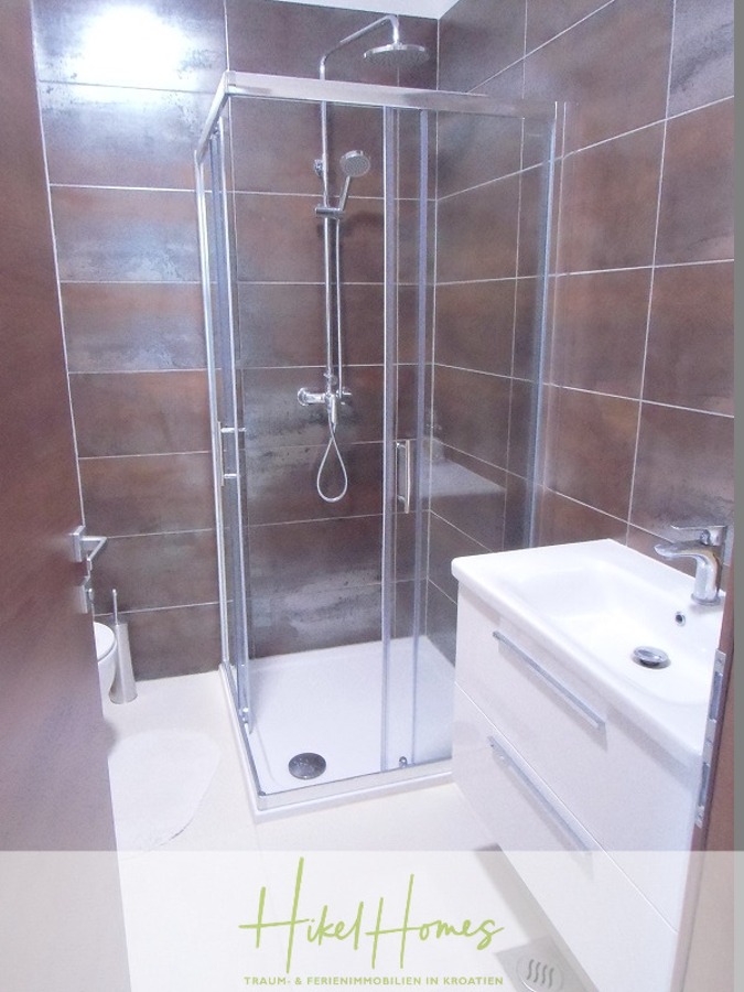 Galerie Wohnung, 3 Schlafzimmer / 70m zum Meer / Pool / 2 Bäder - Modernes Badezimmer mit braunen und weißen Fliesen, einer Glasduschabtrennung, einem an der Wand befestigten Duschkopf, einem kleinen weißen Waschbecken mit Unterschrank, einem Wasserhahn und einer teilweise sichtbaren Toilette. Das "Hikel Homes"-Logo befindet sich am unteren Rand des Bildes. (46579)
