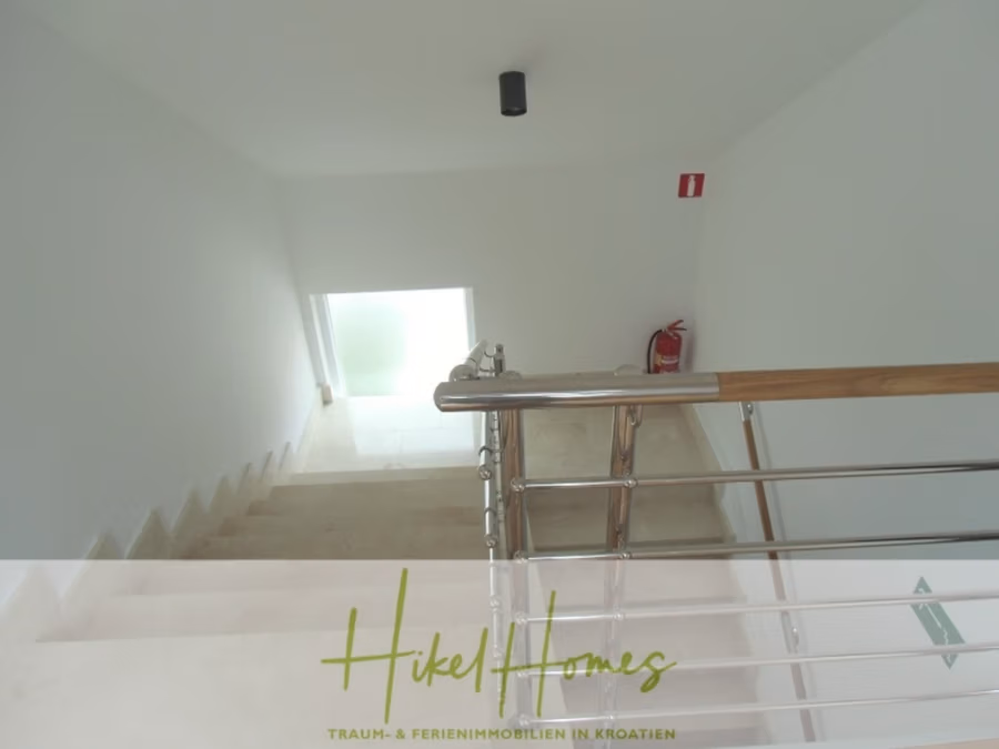 Galerie Wohnung, 3 Schlafzimmer / 70m zum Meer / Pool / 2 Bäder - Eine hell erleuchtete Treppe mit Edelstahlhandlauf führt hinunter zu einer Milchglastür. An der Wand neben der Tür ist ein Feuerlöscher angebracht. Unten im Bild steht der Text „Hikel Homes“ und eine Beschreibung auf Deutsch. (41261)