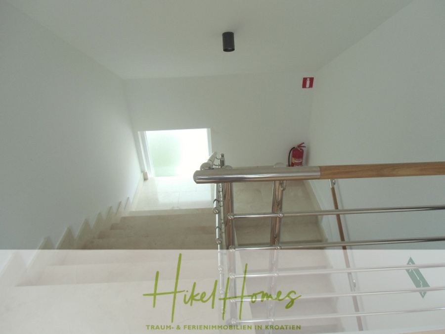 Galerie Wohnung, 3 Schlafzimmer / 70m zum Meer / Pool / 2 Bäder - Ein weißes Treppenhaus mit Metallgeländern führt hinunter zu einer Tür aus Milchglas. Ein roter Feuerlöscher ist an der Wand angebracht, und durch die Tür fällt natürliches Licht ein. Das Logo von Hikel Homes ist am unteren Rand eingeblendet. (46583)
