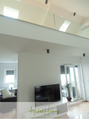 Modernes, helles Wohnzimmer mit einem Fernseher, weißen Wänden, großen Fenstern, einer Balkontür und einer oberen Etage im Loft-Stil mit Glasgeländer. Das Logo von Hikel Homes ist am unteren Rand zu sehen. - Galerie Wohnung, 3 Schlafzimmer / 70m zum Meer / Pool / 2 Bäder