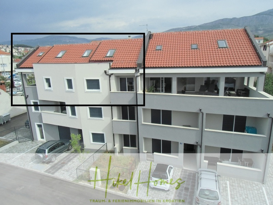 Galerie Wohnung, 3 Schlafzimmer / 70m zum Meer / Pool / 2 Bäder - Ein modernes, hellgraues zweistöckiges Wohnhaus mit Balkonen und einem schrägen Ziegeldach. Im Hintergrund sind die umliegenden Berge und der blaue Himmel zu sehen. Am unteren Rand des Bildes steht "Heikel Homes" geschrieben. (46567)