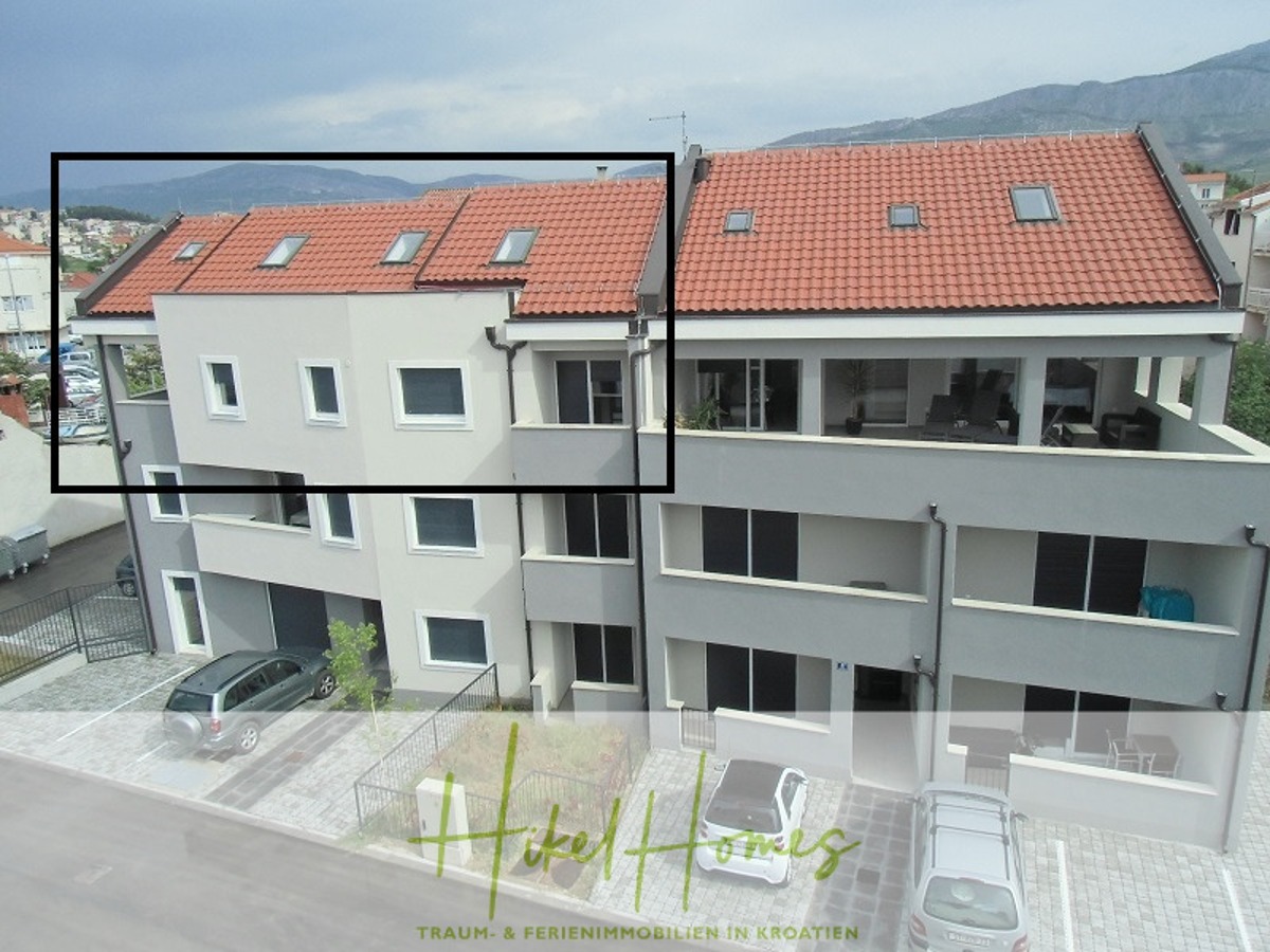 Ein modernes, hellgraues zweistöckiges Wohnhaus mit Balkonen und einem schrägen Ziegeldach. Im Hintergrund sind die umliegenden Berge und der blaue Himmel zu sehen. Am unteren Rand des Bildes steht "Heikel Homes" geschrieben. - Galerie Wohnung, 3 Schlafzimmer / 70m zum Meer / Pool / 2 Bäder