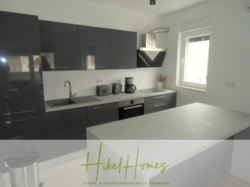 Moderne Küche mit glänzenden dunkelgrauen Schränken, einer Arbeitsplatte mit eingebauter Spüle und Herd, Kleingeräten wie Toaster und Kaffeemaschine und einem Fenster, das Tageslicht hereinlässt. Das "Hikel Homes"-Logo ist am unteren Rand eingeblendet. - Galerie Wohnung, 3 Schlafzimmer / 70m zum Meer / Pool / 2 Bäder