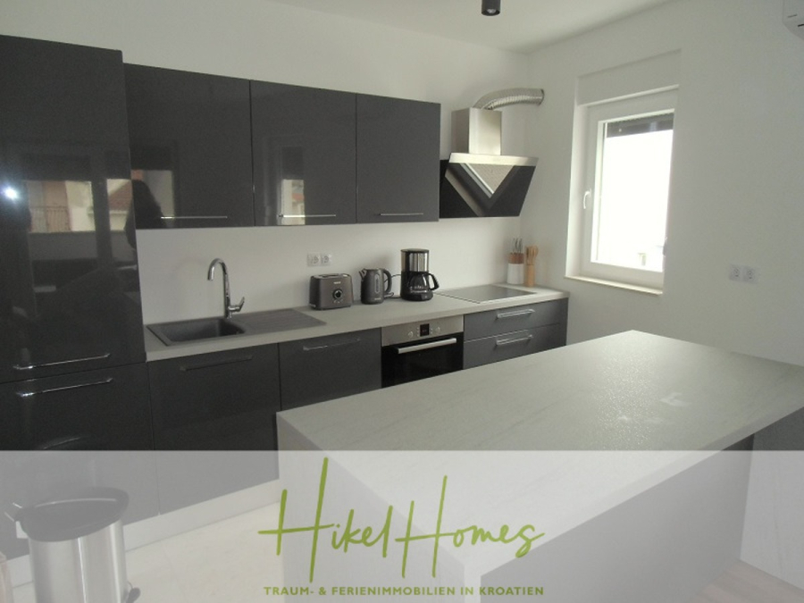Galerie Wohnung, 3 Schlafzimmer / 70m zum Meer / Pool / 2 Bäder - Moderne Küche mit glänzenden dunkelgrauen Schränken, einer Arbeitsplatte mit eingebauter Spüle und Herd, Kleingeräten wie Toaster und Kaffeemaschine und einem Fenster, das Tageslicht hereinlässt. Das "Hikel Homes"-Logo ist am unteren Rand eingeblendet. (46570)