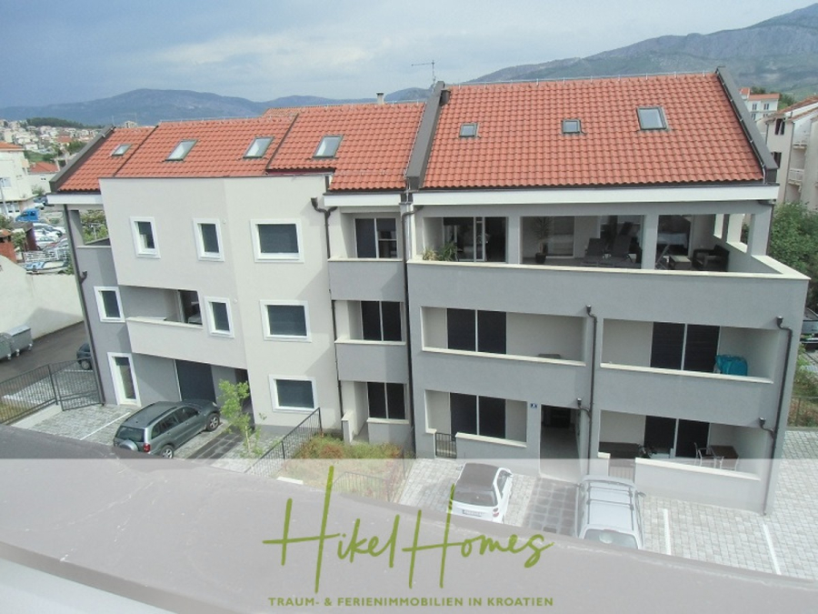 Galerie Wohnung, 3 Schlafzimmer / 70m zum Meer / Pool / 2 Bäder - Ein modernes, dreistöckiges Wohngebäude mit roten Ziegeldächern und weißen Wänden, mit Balkonen und geparkten Autos, umgeben von Bergen im Hintergrund. Das Logo "Hikel Homes" ist am unteren Rand eingeblendet. (46569)