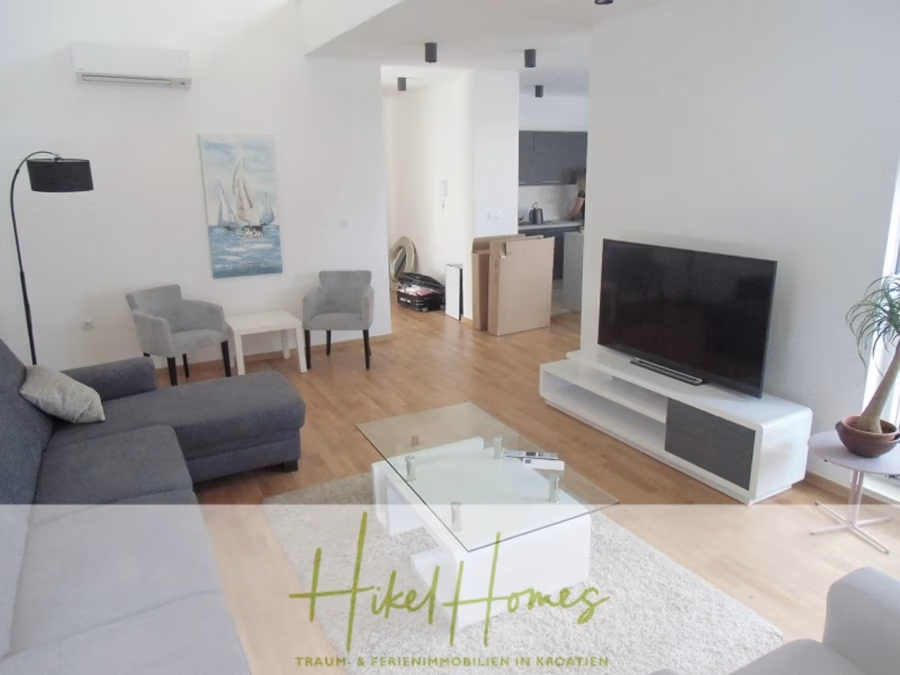 Galerie Wohnung, 3 Schlafzimmer / 70m zum Meer / Pool / 2 Bäder - Modernes Wohnzimmer mit dunkelgrauem Sofa, Glascouchtisch und Flachbildfernseher auf weißem Sockel. Zwei graue Stühle und ein Beistelltisch stehen an der Wand. Der Raum öffnet sich zur Küche mit hellem Holzboden und einem Gemälde eines Segelboots an der Wand. (41255)
