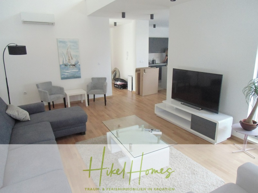 Galerie Wohnung, 3 Schlafzimmer / 70m zum Meer / Pool / 2 Bäder - Modernes Wohnzimmer mit einem grauen Sofa, einem Couchtisch aus Glas, einem Fernseher auf einem weißen Ständer und zwei Sesseln. Der Raum hat einen Holzfußboden und einen offenen Blick in eine Küche. Helle und minimalistische Einrichtung. (46575)