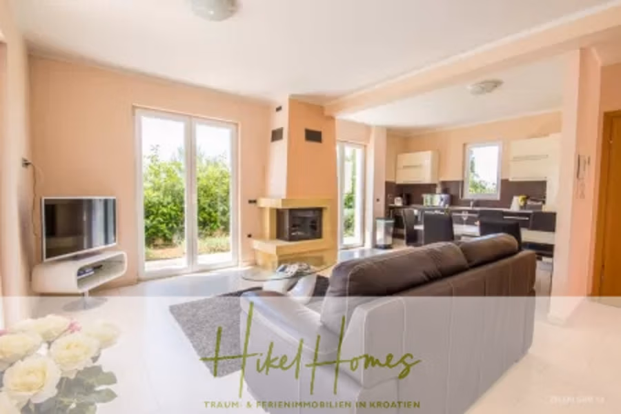 Villa im Mittelmeerstil mit 3 SZ, Pool, Dachterrasse mit Meerblick – möbiliert (15) - Helles Wohnzimmer im modernen Design mit schwarzem Ledersofa, Fernseher und Kamin. Große Fenster bieten Blick ins Grüne. Im Hintergrund befindet sich ein Essbereich, Blumen stehen auf einem weißen Tisch. Der Text auf dem Bild lautet „Hikel Homes“ in grüner Schrift. (41970)
