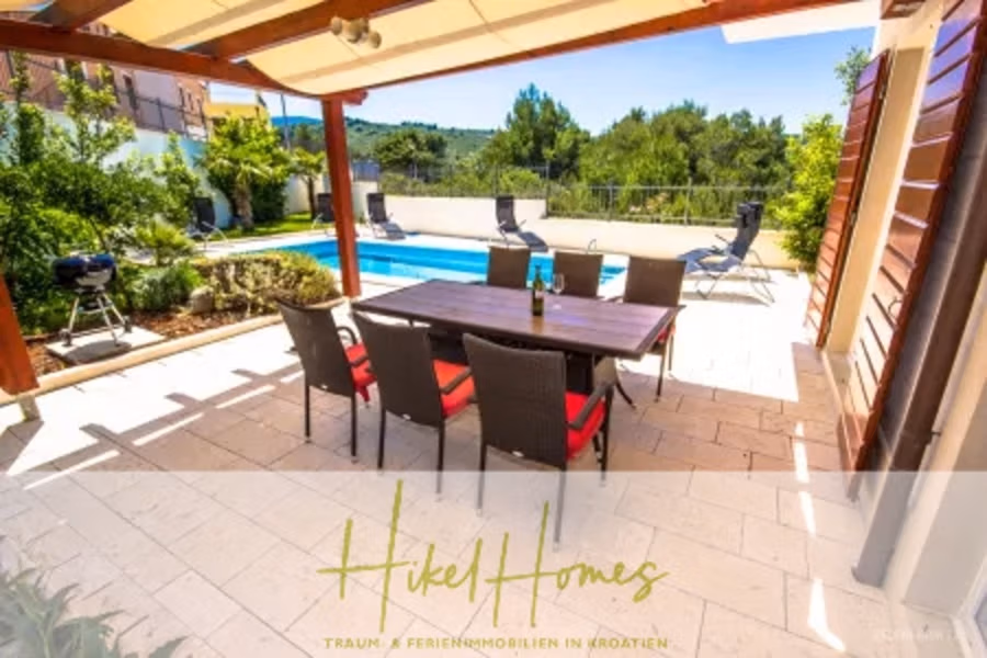 Villa im Mittelmeerstil mit 3 SZ, Pool, Dachterrasse mit Meerblick – möbiliert (15) - Terrasse mit Esstisch und Stühlen unter einer Pergola, in der Nähe eines Pools. Grüne Hügel und Bäume im Hintergrund. Eine Flasche und zwei Gläser stehen auf dem Tisch. Text: „Hikel Homes Traum Ferienimmobilien in Kroatien.“. (41971)
