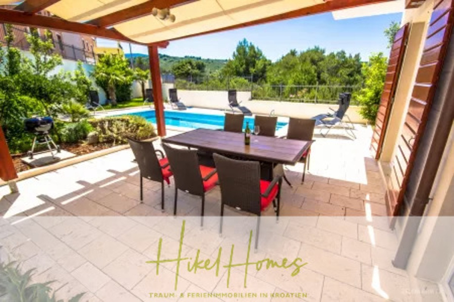 Villa im Mittelmeerstil mit 3 SZ, Pool, Dachterrasse mit Meerblick – möbiliert (15) - Außenterrasse mit einem Esstisch und sechs Stühlen unter einer Pergola, mit Blick auf einen Pool, Sonnenliegen und einen üppigen grünen Garten, mit Hügeln und klarem Himmel im Hintergrund. Logo "Hikel Homes" am unteren Rand. (44855)