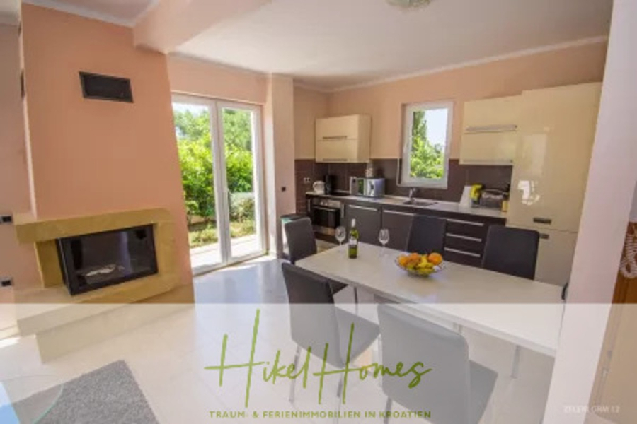 Villa im Mittelmeerstil mit 3 SZ, Pool, Dachterrasse mit Meerblick – möbiliert (15) - Moderne Küche und Essbereich mit hellen Schränken, einem Kamin, einem Esstisch für vier Personen und großen Fenstern mit Blick auf einen grünen Außenbereich. Der Text auf dem Bild lautet "Hikel Homes - Traum- & Ferienimmobilien in Kroatien". (44857)
