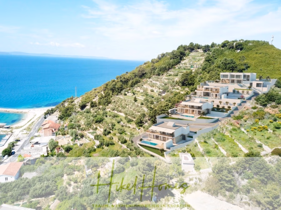 Neubauprojekt Villa Aja – von jedem Zimmer aus Meerblick auf 250m² mit 3 Schlafzimmer / 3 Bädern - Ein Hang mit mehreren modernen, weiß-beigen Villen, umgeben von Grün, überblickt ein Küstengebiet mit Sandstrand und blauem Meer. In Ufernähe befinden sich einige Gebäude mit roten Dächern. Unten ist ein Logo mit Text teilweise sichtbar. (41612)