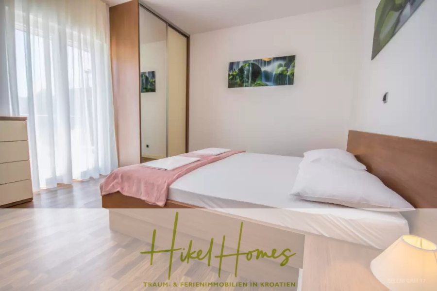 Villa im Mittelmeerstil mit 3 SZ, Pool, Dachterrasse mit Meerblick (3/4) - Ein helles, modernes Schlafzimmer mit einem Doppelbett, weißer Bettwäsche, einer rosafarbenen Decke, einem Kleiderschrank mit Spiegeltüren, einer Kommode, einem Fenster mit durchsichtigen Vorhängen und einem Naturkunstwerk an der Wand. Logo "Hikel Homes" am unteren Rand. (45760)