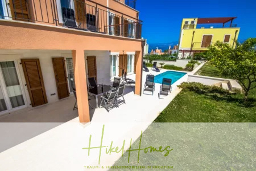 Villa im Mittelmeerstil mit 3 SZ, Pool, Dachterrasse mit Meerblick (3/4) - Ein modernes pfirsichfarbenes Haus mit hölzernen Fensterläden, einer überdachten Terrasse mit einem Essplatz im Freien, Liegestühlen, einem Swimmingpool und einem grünen Rasen unter einem klaren blauen Himmel. Der überlagerte Text lautet "Hikel Homes". (45744)