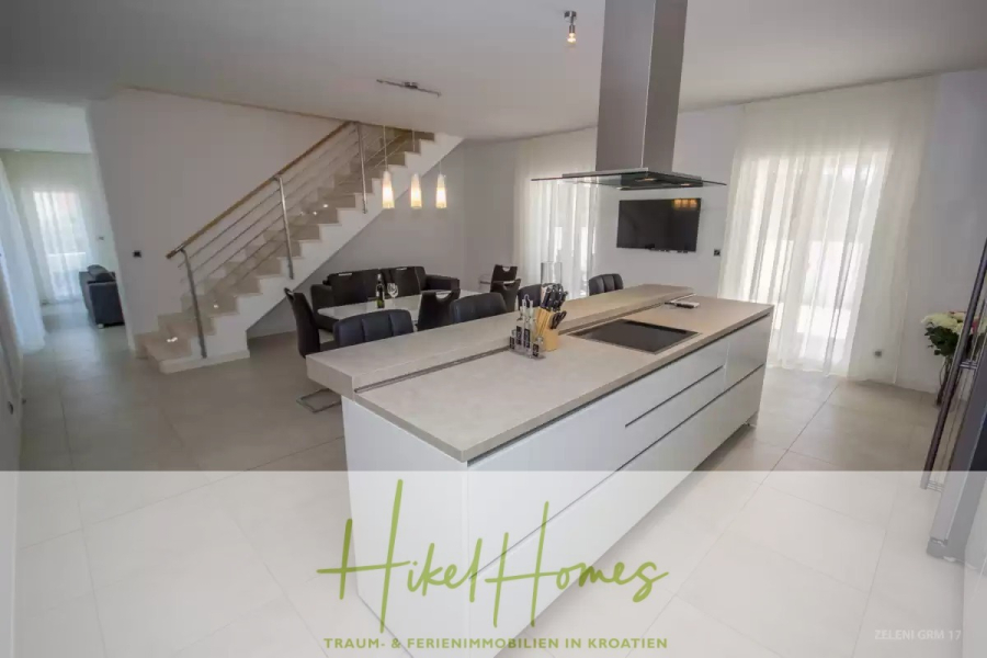 Villa im Mittelmeerstil mit 3 SZ, Pool, Dachterrasse mit Meerblick (3/4) - Moderne, offene Küche und Essbereich mit großer weißer Insel, Einbauherd und Dunstabzugshaube. Eine Treppe mit einem Glasgeländer führt nach oben. Der Raum ist hell mit natürlichem Licht durch transparente Vorhänge. (45763)