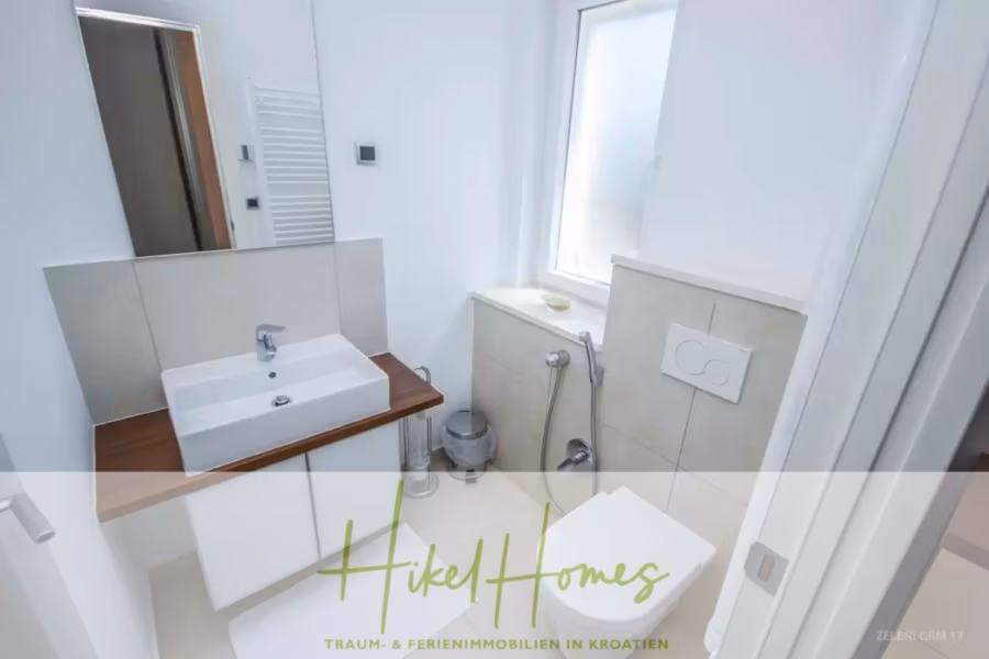 Villa im Mittelmeerstil mit 3 SZ, Pool, Dachterrasse mit Meerblick (3/4) - Ein kompaktes, modernes Badezimmer mit weißen Wänden und Fliesen. Es verfügt über ein quadratisches Waschbecken mit Holzplatte, eine wandmontierte Toilette und ein kleines Fenster. Das Logo „Hikel Homes“ ist unten im Bild eingeblendet. (43311)