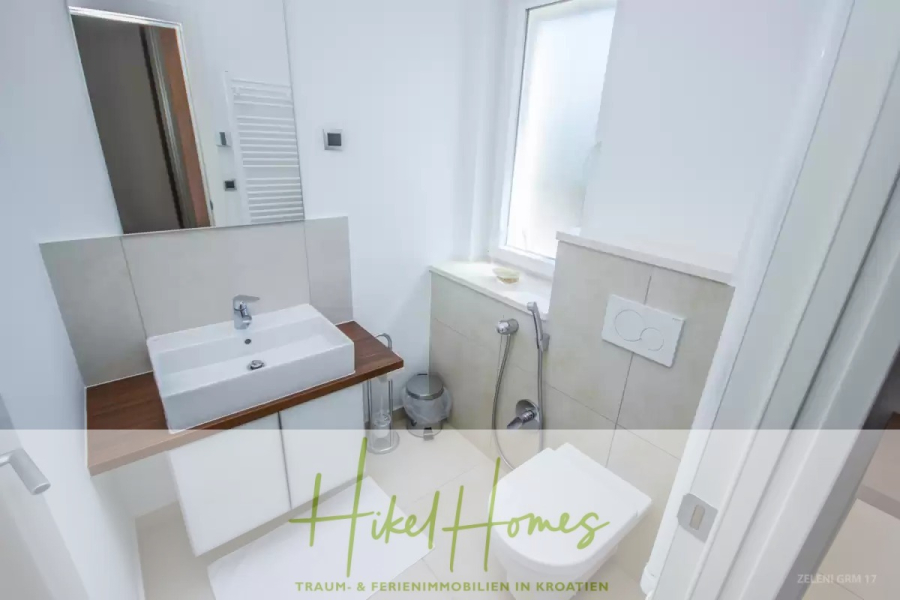 Villa im Mittelmeerstil mit 3 SZ, Pool, Dachterrasse mit Meerblick (3/4) - Modernes, kompaktes Badezimmer mit weißem Waschbecken auf einem Holztisch, Spiegel, Toilette, Handbrause, Fenster und neutralen Fliesen. Am unteren Rand des Bildes ist der Schriftzug "Hikel Homes" zu sehen. (45751)